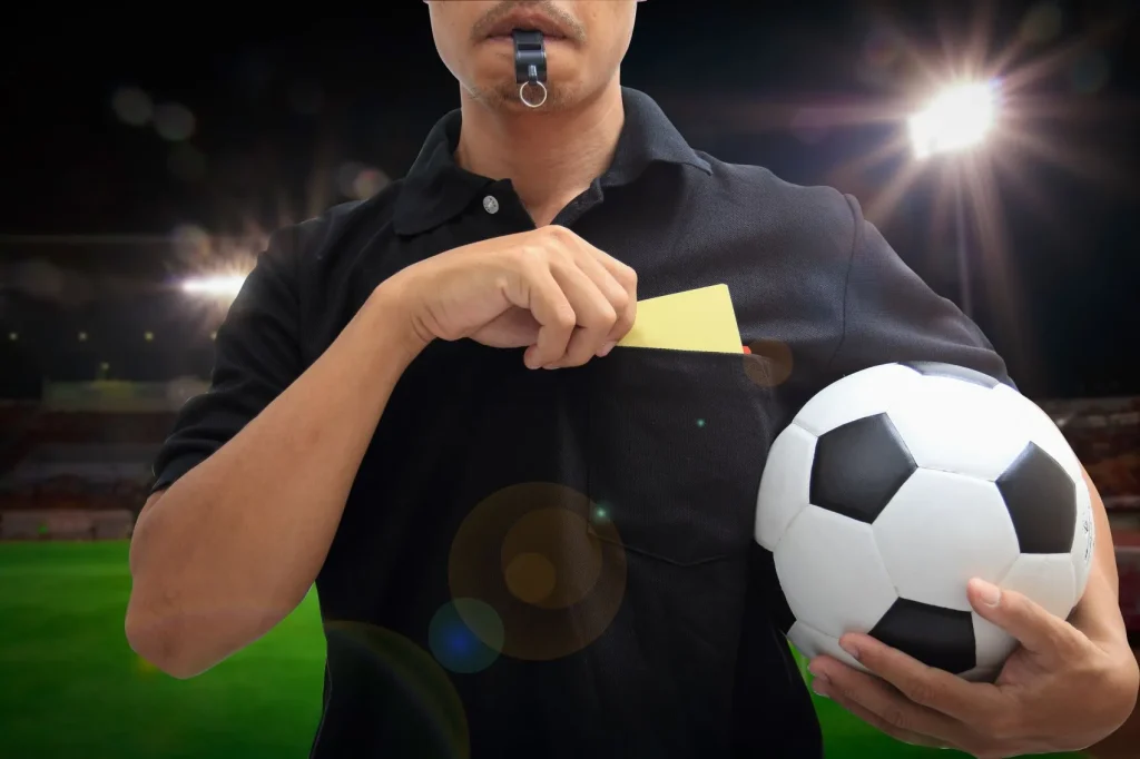 Schiedsrichter hält einen Fußball in einer Hand, eine Pfeife im Mund und holt mit der anderen Hand gerade eine Gelbe Karte heraus