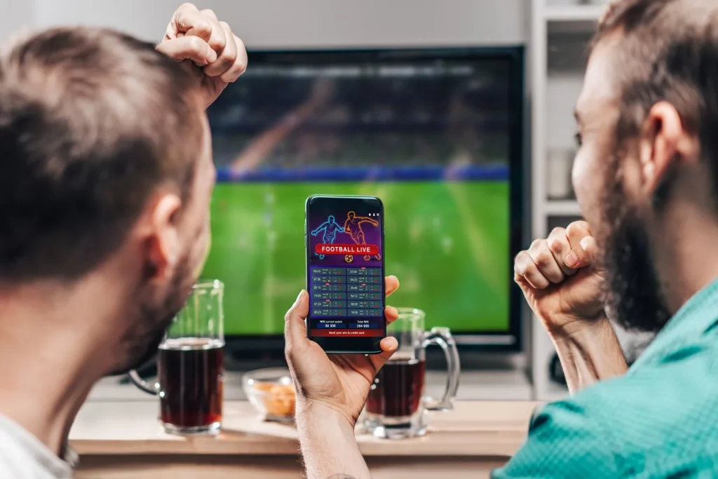 Zwei Männer schauen während eines Fußballspiels auf dem Smartphone, wie ihre Sportwetten verlaufen