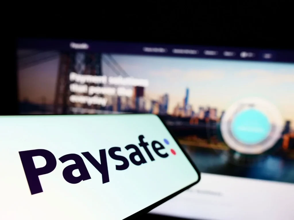 Logo von Paysafe auf einem Smartphone, positioniert vor einem Laptop mit der Paysafecard-Website