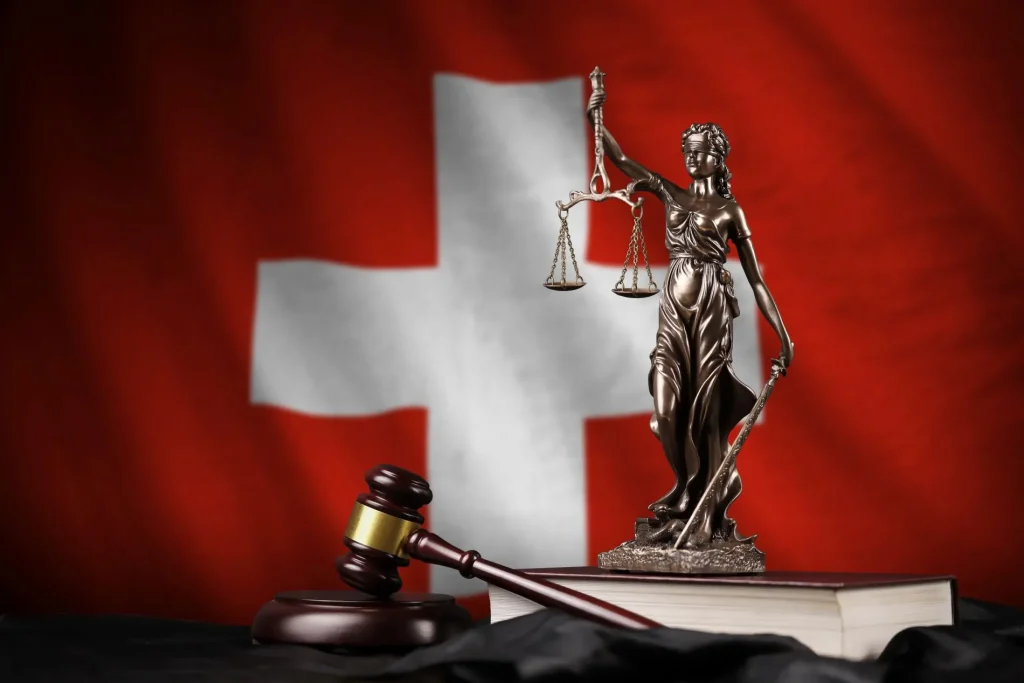 Justizia-Figur mit einer Waage in der linken Hand, zu ihren Füßen ein Richterhammer und um Hintergrund die Landesflagge der Schweiz